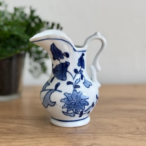 May include: A small, white ceramic pitcher with a blue floral design. The pitcher has a curved handle and a spout. The floral pattern includes stylized flowers and leaves. The pitcher is approximately 5 inches tall.