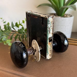 May include: A vintage mortise door lock set featuring two glossy black porcelain knobs attached to a rectangular metal box mechanism. The hardware includes a white circular mounting plate and a central metal spindle, showing signs of wear.
