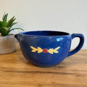 May include: A navy blue ceramic mixing bowl with a handle and spout. The bowl is decorated with yellow leaf designs and a red dot. A small potted plant is visible in the background.