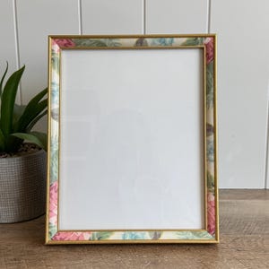 May include: A rectangular picture frame with a thin gold metallic border and an inner inlay featuring a floral pattern in shades of pink, green, and blue. The frame surrounds a blank white rectangular center area, designed for displaying photos.