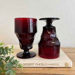 May include: Two ruby red vintage goblets with a geometric design. One goblet is upright, the other is upside down. The goblets are on a book with the text "A Desert Pueblo Harte".