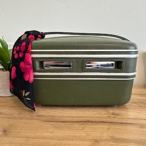 May include: Olive green vintage suitcase with white trim and chrome accents. A black scarf with a red floral pattern is draped over the handle. The suitcase has a handle and two chrome latches.