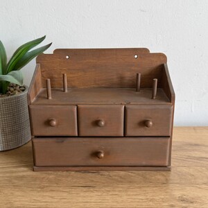 May include: A wooden sewing organizer with three small drawers and a larger bottom drawer. The organizer has a shelf with wooden pegs and is stained a warm brown color. The piece is designed to hold sewing supplies.