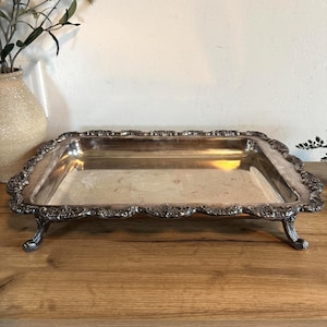 May include: A rectangular silver-colored serving tray with ornate detailing around the edges and on the feet. The tray is set on a wooden surface, with a vase and greenery in the background.