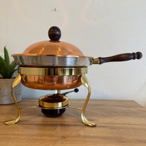 May include: A copper and brass chafing dish with a wooden handle and a dark wooden knob on the lid. The dish has a gold-colored stand and a small burner below. The chafing dish is designed for keeping food warm.