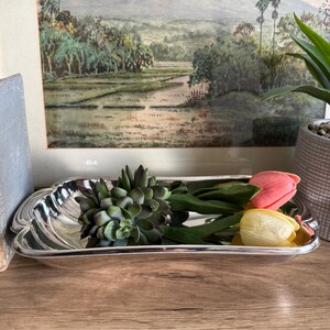 Op de afbeelding: Een zilverkleurige, rechthoekige schaal met een decoratieve rand bevat een groene vetplant en twee tulpen, &eacute;&eacute;n roze en &eacute;&eacute;n geel. De schaal staat op een houten oppervlak, met een ingelijst landschapsschilderij op de achtergrond.