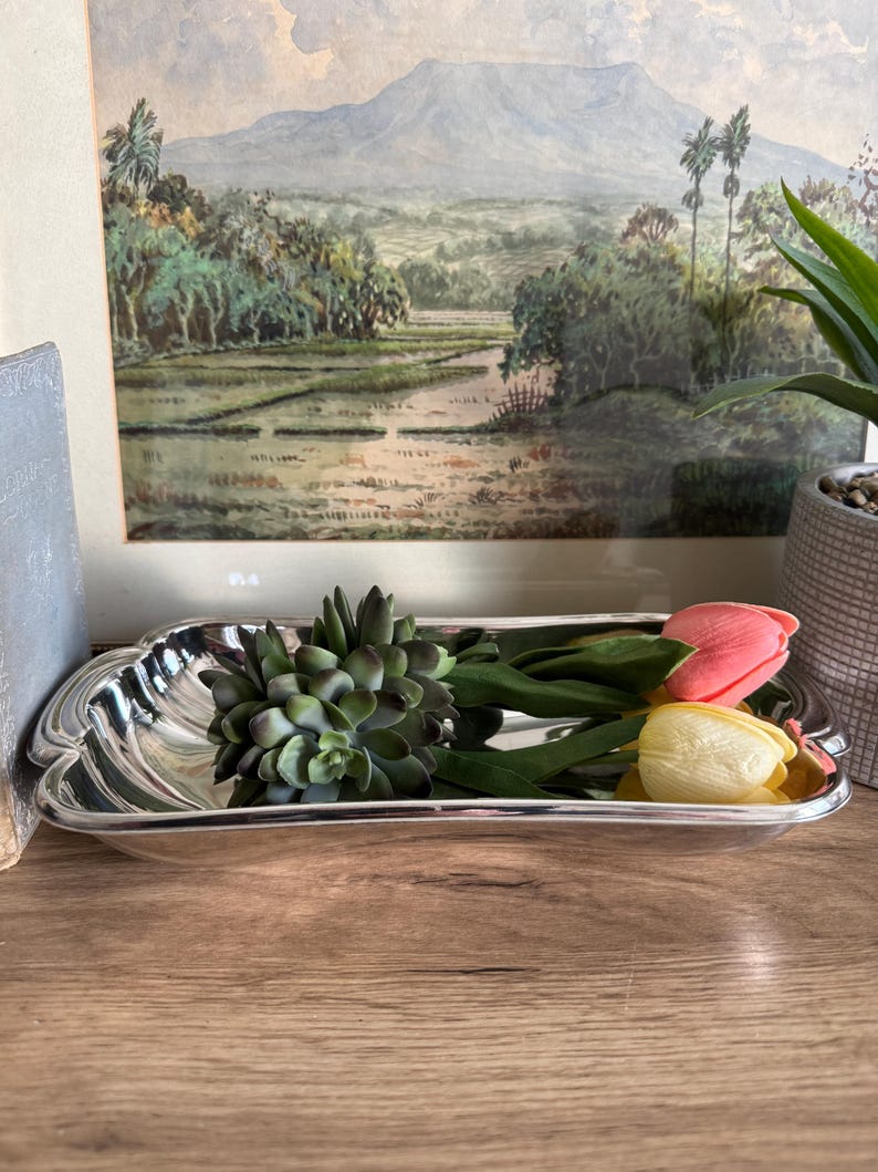 Op de afbeelding: Een zilverkleurige, rechthoekige schaal met een decoratieve rand bevat een groene vetplant en twee tulpen, &eacute;&eacute;n roze en &eacute;&eacute;n geel. De schaal staat op een houten oppervlak, met een ingelijst landschapsschilderij op de achtergrond.