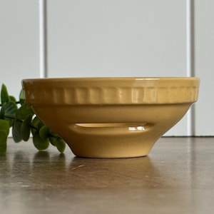May include: A mustard yellow ceramic bowl featuring a wide horizontal cutout near the base and a decorative fluted or ribbed pattern along the upper rim. The bowl has a smooth, solid-colored finish and a wide, open top design.