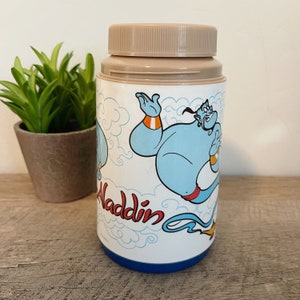 May include: A white plastic Aladdin themed lunchbox with a blue genie character on it. The genie is flying and has a blue body, orange arms, and a red sash. The lunchbox has a brown lid and a blue bottom.