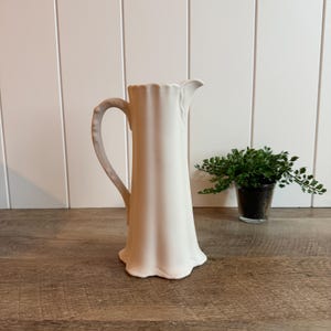 May include: A tall, matte white ceramic pitcher featuring a fluted, vertical ribbed design along its body, a curved handle with a textured grip, and a flared base. The pitcher has a distinct pouring spout at the top rim.