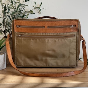 May include: A vintage briefcase with a brown leather top and handle, and a khaki canvas body. The bag has multiple zippered compartments and a long, adjustable leather shoulder strap. The bag is sitting on a wooden surface.