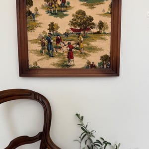 May include: A framed vintage print depicting a golf scene with figures in period attire. The artwork features a brown wooden frame and a beige background with green and brown trees and figures. The print is mounted on a white wall.