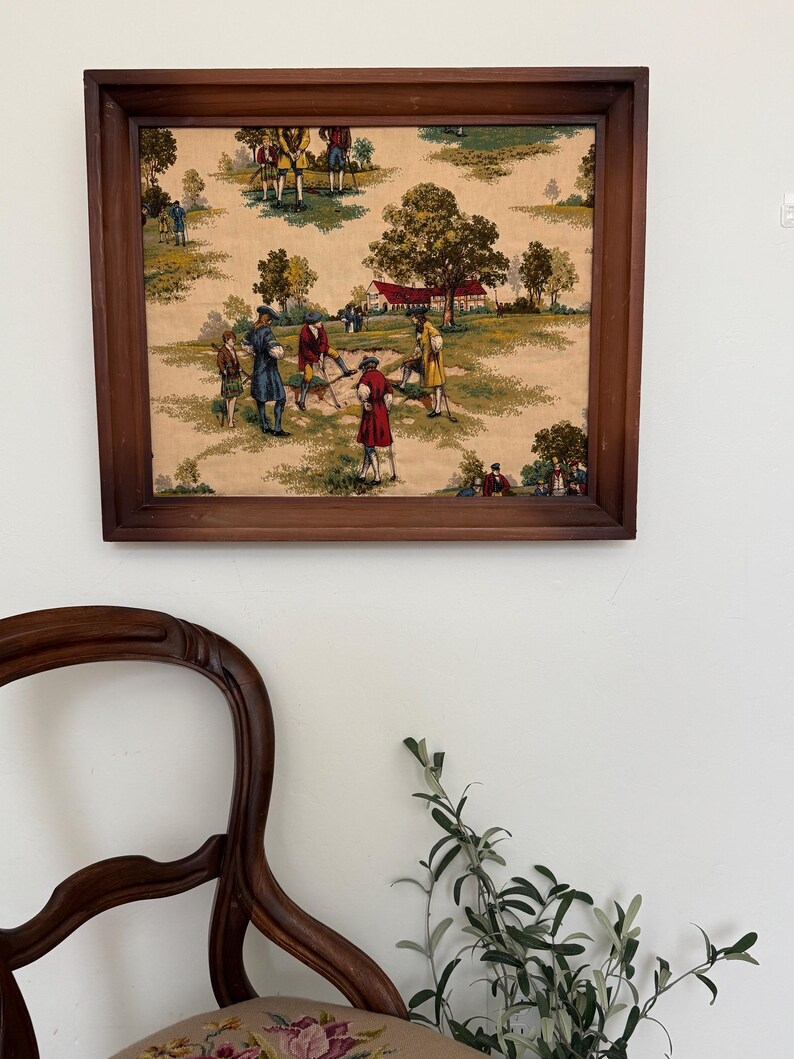 May include: A framed vintage print depicting a golf scene with figures in period attire. The artwork features a brown wooden frame and a beige background with green and brown trees and figures. The print is mounted on a white wall.
