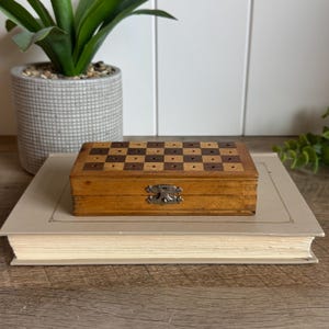 May include: A rectangular wooden box featuring a checkerboard pattern of light and dark wood squares on the lid, each with a small hole. The box has a silver-colored metal latch on the front and is resting on top of a closed, light-colored book.