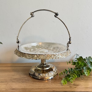 May include: A silver-toned, two-tiered serving stand with an ornate floral design on the plate and base. The stand has a curved handle and sits on a wooden surface. The overall design is vintage and elegant.