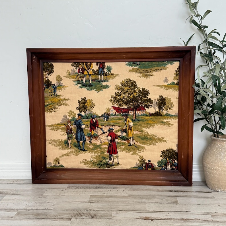 May include: A framed fabric panel featuring a pastoral scene with figures in period clothing playing golf. The artwork is set in a brown wooden frame. The fabric has a beige background with green, red, yellow, and blue accents.