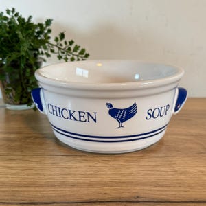 May include: White ceramic chicken soup bowl with blue accents. The bowl has two handles and the words "CHICKEN SOUP" in blue, along with a blue chicken illustration. The bowl is sitting on a wooden surface.