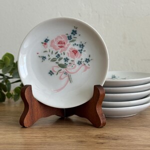 May include: White ceramic plate with a pink rose bouquet design, displayed on a wooden stand. The bouquet features pink roses, blue accents, and a pink ribbon. Additional plates are stacked in the background.