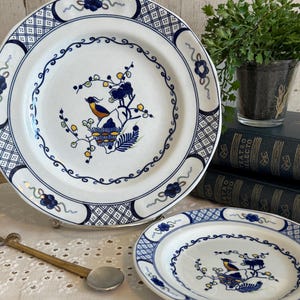 May include: White ceramic plates with blue floral and bird designs. The plates have a blue border with a repeating pattern. A smaller plate is also visible. The plates are part of a dinnerware set.