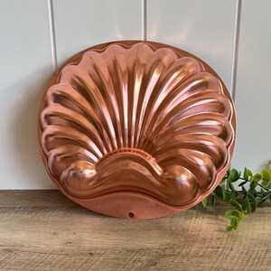 May include: A round copper-colored baking mold featuring a raised, embossed seashell design with radiating ridges. The metallic surface has a polished finish with a small circular hole at the bottom edge for hanging, showcasing a classic scallop shell shape.