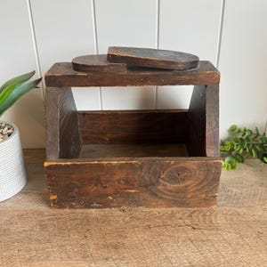 May include: A dark brown wooden caddy with a rectangular base, angled side panels, and a top handle. The wood features a visible grain pattern with a weathered finish, showing natural knots and variations in the dark brown stain throughout the structure.