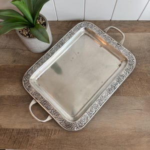 May include: A rectangular silver-toned metal serving tray featuring a raised border with a detailed embossed grape and leaf pattern. The tray includes two curved handles on opposite ends and a smooth, reflective central surface area.