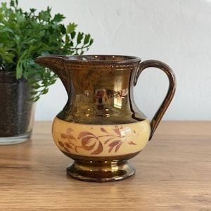 May include: A small, decorative pitcher with a brown and gold metallic glaze. The pitcher features a cream-colored band with a floral design. The handle and spout are brown. The pitcher is likely made of ceramic or porcelain.