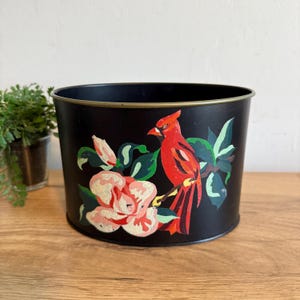 May include: Black metal planter with a gold rim. The planter is decorated with a hand-painted design of a red cardinal bird perched on a branch with pink and white flowers and green leaves.
