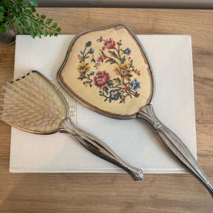 May include: A vintage vanity set featuring a hand mirror and a brush. The mirror has a floral needlepoint design in red, yellow, and blue. Both items have silver-toned handles. The brush has soft, light-colored bristles.