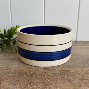 May include: A round ceramic bowl with a cream-colored exterior featuring a wide horizontal dark blue band and two thin black stripes. The interior of the bowl is a solid dark blue. The ceramic surface has a subtle crackle glaze finish.