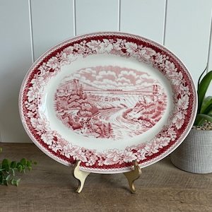 May include: An oval ceramic platter featuring a deep red transferware pattern. The center depicts a landscape with a suspension bridge over a river, while the wide rim is decorated with a detailed border of embossed-style leaves and floral motifs.