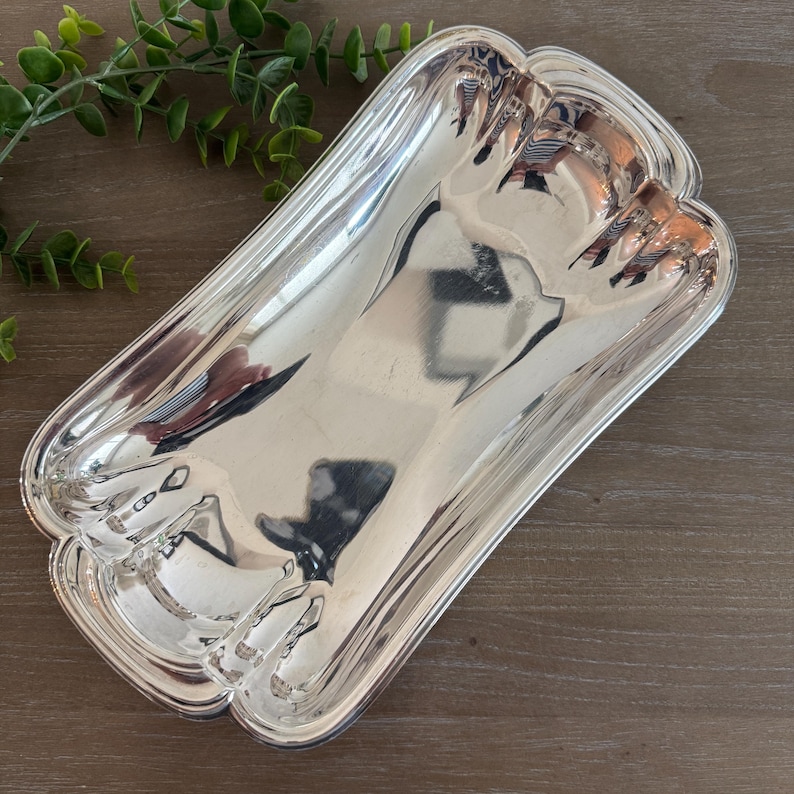 Op de afbeelding: Een rechthoekige, zilverkleurige serveerschaal met een geschulpte rand. De schaal heeft een reflecterend oppervlak en de randen zijn iets verhoogd. De schaal is leeg en ligt op een houten oppervlak.