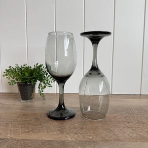 May include: Two wine glasses made of translucent gray glass. One glass stands upright on its circular base, while the other is inverted, resting on its rim. The glass features a smooth, curved bowl, a slender stem, and a wide, stable base.