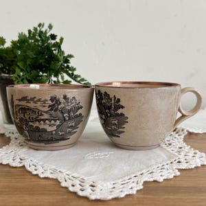May include: Two beige ceramic teacups with a brown rim and handle. Each cup features a black and white pastoral scene with trees, a bridge, and grazing animals. The cups are placed on a white lace doily.