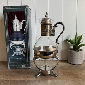 May include: A clear glass carafe with a silver-plated metal collar, lid, and ornate handle. The carafe rests on a matching silver-plated metal stand with three curved legs and a central base for a tea light candle. The original box is included.