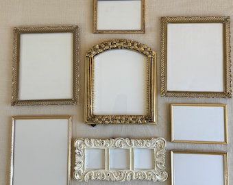 May include: An assortment of ornate picture frames in various shapes and sizes. The frames are primarily gold and cream-colored, with intricate detailing and empty white spaces for photos or artwork. The collection includes rectangular, square, and arched frames.