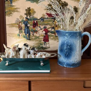 May include: A blue ceramic pitcher with a floral design and a handle, next to a silver tray holding small dog figurines. A green book and a framed tapestry with figures in period clothing are also visible.