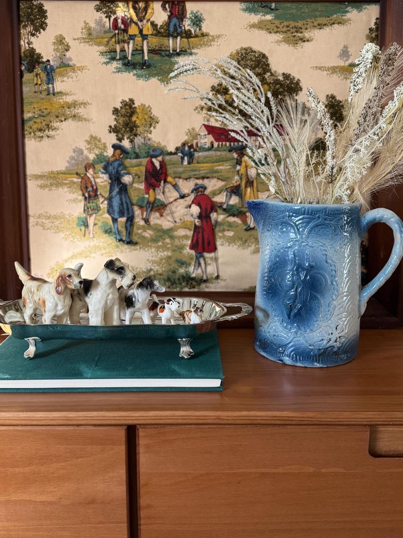 May include: A blue ceramic pitcher with a floral design and a handle, next to a silver tray holding small dog figurines. A green book and a framed tapestry with figures in period clothing are also visible.