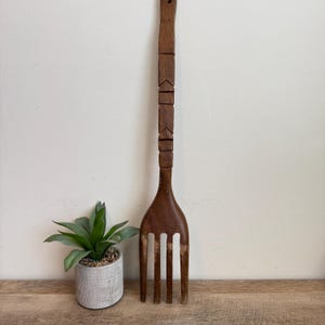 May include: A large, vintage wooden fork with a long handle and four tines. The handle features carved geometric designs. The fork is a dark brown color and appears to be handmade. It is displayed next to a small potted plant.
