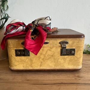 May include: A vintage suitcase with a yellow textured exterior and a brown top. A red and white patterned scarf is draped over the top. The suitcase has metal latches and a leather handle.