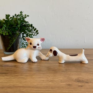 May include: Two ceramic figurines: a cat and a dog. The cat is cream-colored with pink ears and brown details. The dog is also cream-colored with brown spots. Both are on a wooden surface.