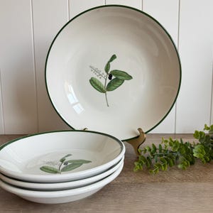 May include: A set of white ceramic bowls featuring a dark green rim and a botanical illustration of green leaves and small black berries in the center. One bowl is displayed upright on a stand, while three others are stacked in the foreground.