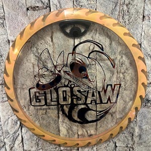 May include: A clear disc golf disc with a cartoon bee graphic and the word "GLOSAW" in black. The disc has a gold-colored, saw-tooth edge and a black center piece. The text "LE FUZED BUZZZ" is below the word "GLOSAW".