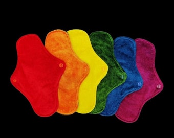 May include: A collection of reusable cloth menstrual pads in a rainbow of colors: red, orange, yellow, green, blue, and purple. Each pad has a curved shape and a snap closure. The pads are arranged against a black background.