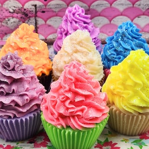 May include: A collection of six colourful cupcake soaps with glitter frosting. The cupcakes are in shades of purple, pink, yellow, blue, orange and white.