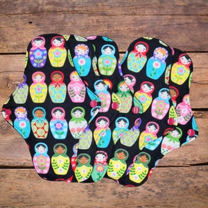 May include: A set of five black reusable cloth pads featuring a colourful Russian nesting doll pattern. The dolls are wearing traditional clothing and have flowers on their dresses.