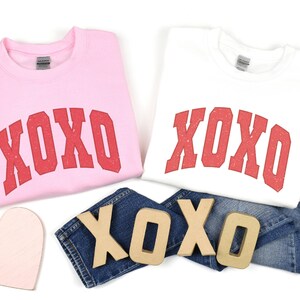 May include: Two sweatshirts with the word "XOXO" printed in red on the front. The sweatshirts are pink and white. There are also wooden letters spelling "XOXO" on a pair of blue jeans.