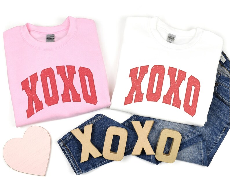 May include: Two sweatshirts with the word "XOXO" printed in red on the front. The sweatshirts are pink and white. There are also wooden letters spelling "XOXO" on a pair of blue jeans.