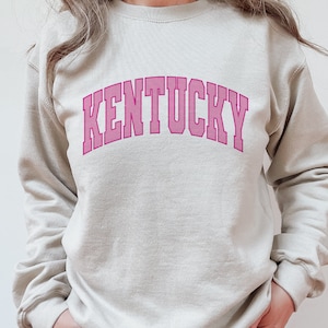 Pode incluir: Uma camisa de moletom bege claro com um design de arco rosa que diz "KENTUCKY".
