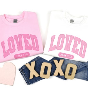 Op de afbeelding: Twee roze sweatshirts met het woord "LOVED" in roze letters en de tekst "JOHN 3:16" eronder. De sweatshirts liggen op een witte achtergrond met een roze hart en houten letters die "XOXO" spellen.
